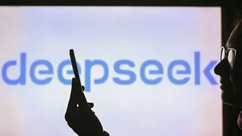 Getty Images A woman holds a cell phone in front of a computer screen displaying the DeepSeek logo