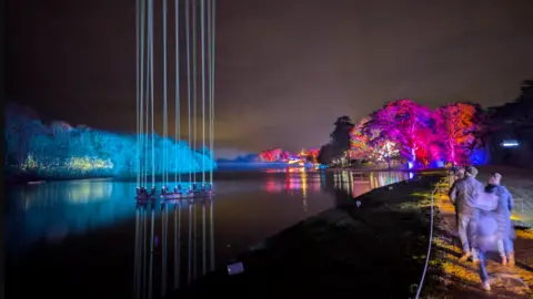 Lucie Johnson A lake with columns of vertical light shining up from the water. In the distance you can see trees and buildings decorated with bright Christmas colourful lights. A family walks along a path next to the lake.