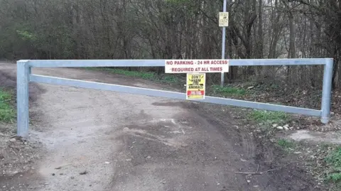 North Northamptonshire Council A large metal gate at Ecton that is closed. A lamp post is behind it, with a row of trees. There are signs on the gate. 