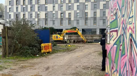 Heavy machinery and a police officer are at the site. A grey building with lots of windows is at the site. A brown fence and a bush are at the left of the image. A small yellow plastic barrier is next to the bush. 