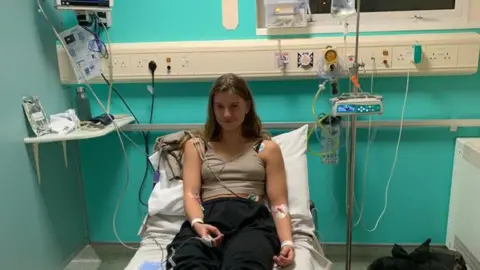 Beth Edwards Beth lies on a hospital bed in a clinical room with teal walls, connected to medical monitoring equipment and IV lines. Medical devices, cables and supplies are positioned around the bed, with personal belongings on the floor nearby.