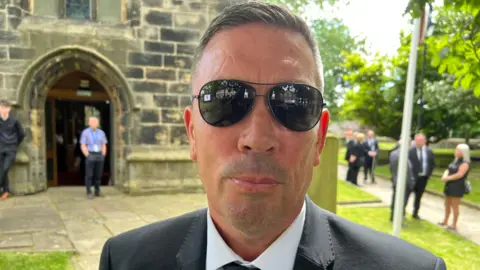 Paul Ogden/BBC A man with short grey hair wearing black sunglasses looks at the lens. Behind him is a church and other people dressed in black. He has a black suit jacket on.