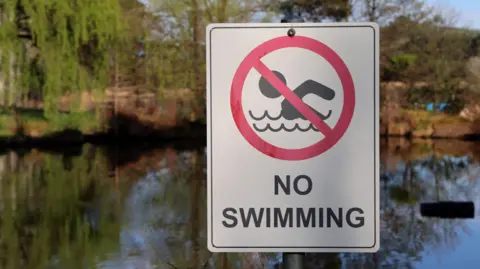 A white sign is in the foreground, which reads NO SWIMMING. There is a picture of someone appearing to swim, with a red line through the image. In the background is a pond area and trees.