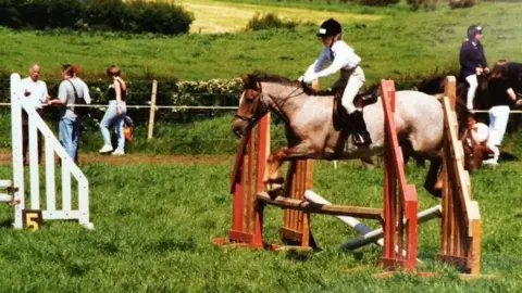 Harry Hall white riding gear and a young jockey in a black helmet, riding a brown horse because it jumps on red wooden structures on a green lawn, there are spectators in the background