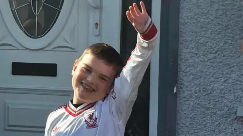 Dáithí Mac Gabhann. He is smiling and holding his hand up in the air. He is wearing a white long sleeve Liverpool jersey. A white door is behind him.