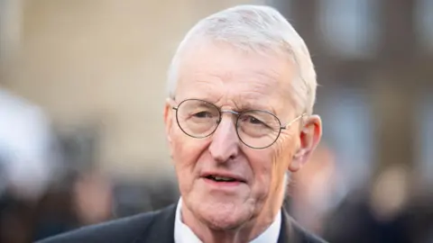 Hilary Benn has short grey hair and thin black frames glasses and is wearing a dark suit.