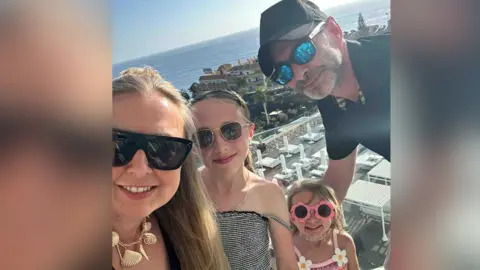 Group photo of the Rimmer Family abroad. All wear sunglasses smiling. A couple in their late thirties to early forties pose with their two young daughters for the selfie.