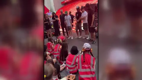 Anfield Road Primary School The children sit on the floor at Anfield Stadium looking at the band members stood in front of them.