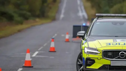 A police car is parked within a coned-off section of a single carriageway road.
