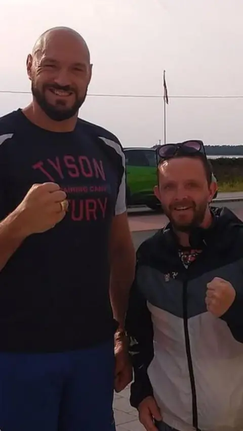 RICHARD HORNE Tyson Fury wears a t-shirt that reads Tyson Fury, he is bald and very tall, he stands next to Richard who has brown stubble and sunglasses on his head, they both smile brightly posing with clenched fists.