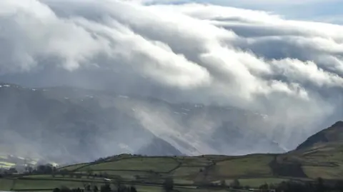 Weather Watchers / IcyMike Fields astride a valley, with mountains in the background and lowering clouds above. It appears to be raining or snowing in the distance.