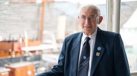 Art Lewry/Culture Communications Collective Gibbie Fraser smiling straight at the camera. He is wearing a blue blazer with coloured pins in the lapel. He has white hair and clear glasses.