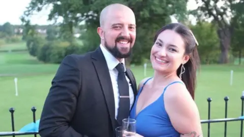 Joe Mintz and Vanessa Curto stand in front of black railings with a golf putting green and trees behind them. Joe is wearing a black suit, white shirt and black tie, has a shaved head and dark beard. Vanessa wears a blue dress, has a piece of snake jewellery on her upper arm, and has long brown hair tied back. Both are holding champagne flutes and are smiling at the camera.