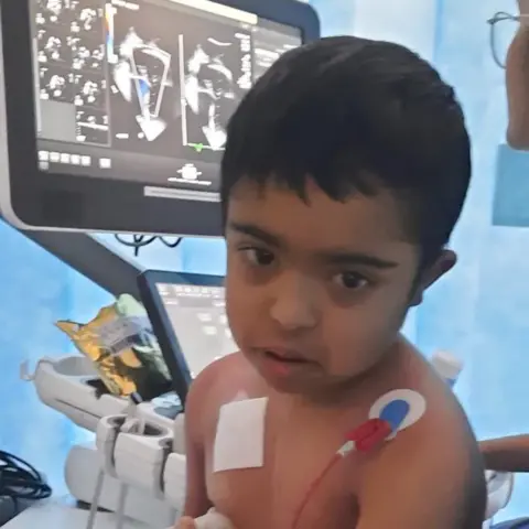 Family handout A boy with dark hair is sitting upright in a hospital bed with tubes across his body. A medic is using an ultrasound on his chest. 
