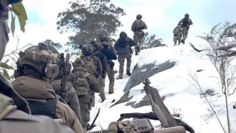 Victoria Police Officers in dark brown trek up a snowy, steep and rocky hillside in Victoria.