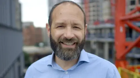 Back the Bakerloo Kieron Williams is seen with a short beard and is smiling at the camera while wearing a light blue button-up shirt. He is standing outdoors with a background featuring a construction site, including tall buildings, scaffolding, and a red crane.