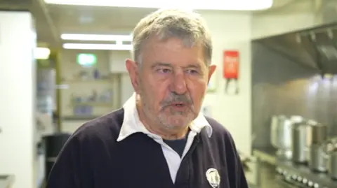 Bill Ashton standing inside a kitchen. He is wearing a dark purple rugby shirt and is looking directly at the camera.