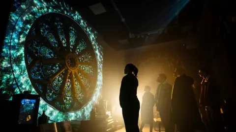 Visitors to The Inverness Castle Experience stand looking at an exhibit called the Rose Window. The visitors are bathed in light from the stained glass window. 