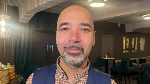 A man with a shaven head inside an art deco cinema wearing a orange checked shirt with a blue waist coat 