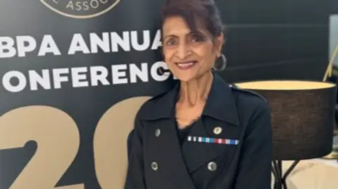 A woman wearing a black blazer jacket and black trousers stands in front of a large black board with text on it which reads "NBPA Annual Conference 2024". She has several embroidered medal ribbons on her jacket.