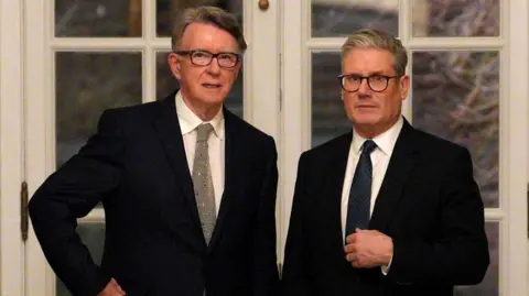 PA Un Mandelson y Starmer serios están uno al lado del otro con trajes negros.