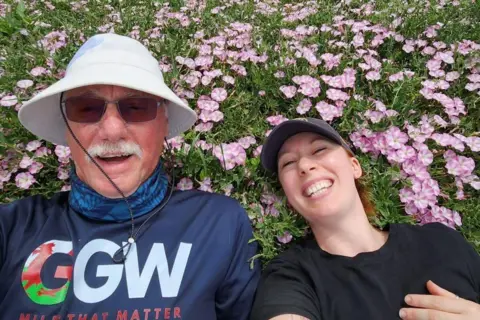 Kyla Norrie John and Kyla smile for a selfie as they lay in a flower bed for a rest during their walking challenge. The purple flowers surround their heads, they both wear sunhats. 