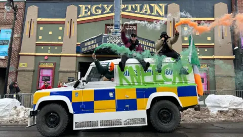 James Clarke Kneecap members on top of a white land rover, modified to look like a PSNI vehicle, with Kneecap graffiti... two bandmates are sat on the roof letting off green white and orange flares as they pull up to a deco-style Egyptian cinema in Utah, signs advertising Sundance Film Festival are visible and the ground has some snow on it 