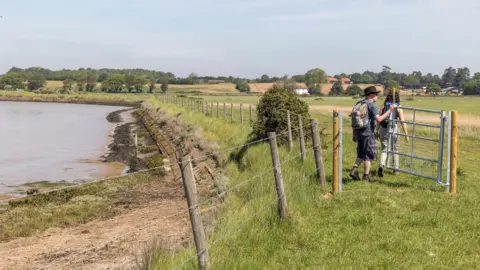 Two walkers walk through a gate that connects two fields. There is a river on the left hand side. In the distance there are some buildings and further fields. 