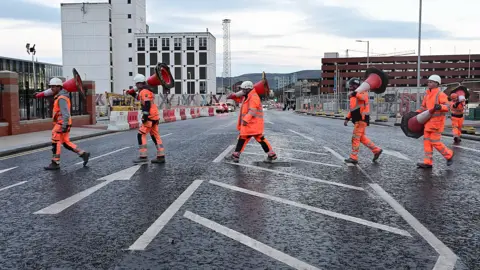 Pacemaker A group of people wearing orange high visibility clothes moving bollards off the road. 