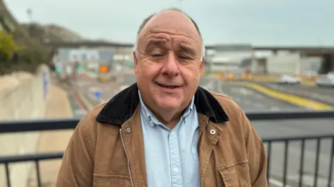 A man in a shirt and brown jacket. The entrance to the Port of Dover can be seen in the background.