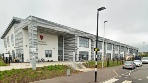 Google A general view of Paradykes Primary School. It shows a modern building which is mainly white, with grey, white and dark grey cladding around the sides and windows, which stretch down the length of the image. A street light, which is mainly black with white bands on it is in the foreground of the image.