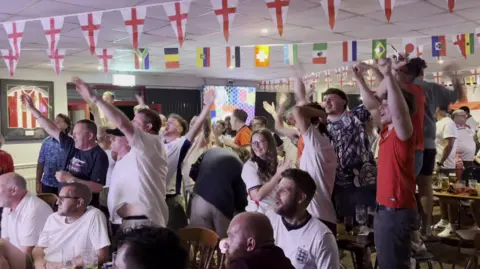 BBC Football fans singing and pointing inside a large room with lots of England flags hung on the ceiling