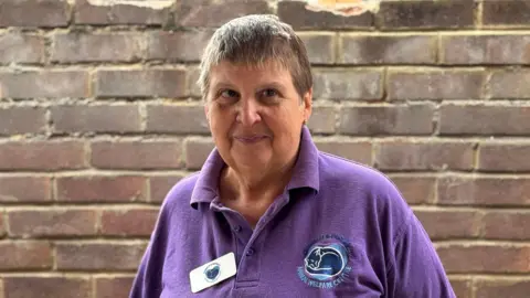 George King/BBC Suzanne Tripp standing in front of a brick wall wearing a purple polo shirt with the charity's logo on it. She is looking into the camera.