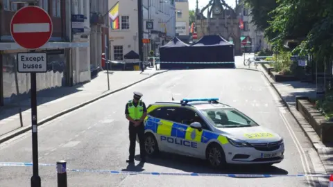 EDDIE MITCHELL A police officer in front of a police car at the scene of the incident. There is police tape at both ends of a street and a large blue police tent in the distance. 