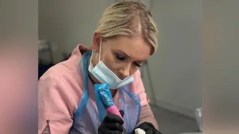 Kirsty Jackson is pictured using a tattoo gun. She wears a blue mask on her face, black gloves and pink uniform with a blue plastic overall over the top. Her blonde hair has been tied back.