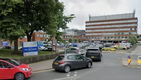 Google A queue of cars turning into a car park. A sign at the entrance says York Hospital. Further away is a large tall building which says York Hospital Main Entrance on the front.
