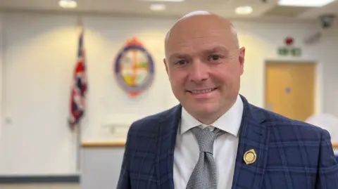 Andy Barnes smiling at the camera. He has a bald head and is wearing a blue tartan suit with a gold circular badge on his lapel. He is wearing a grey tie over a white shirt. Behind him is a union flag and coat of arms out of focus.
