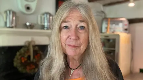 JOANNA TAYLOR/BBC Susan Elliott looking at the camera. Behind her is the interior of her cottage including a fireplace and mantlepiece, which is out of focus. 