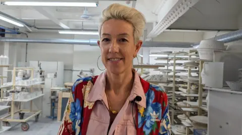Emily wears a colourful jacket over pink overalls, she is wearing hoop earrings and has bright blonde, short hair with a quiff. She is on the factory floor surrounded by shelves displaying mugs and plates, and looking directly at the camera. 