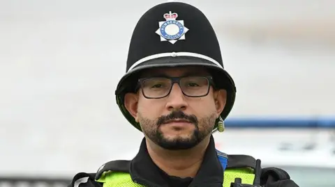 PC Mourad Karaouani in his Humberside Police uniform with helmet, radio and body-worn camera. He has black hair and beard and wears glasses.