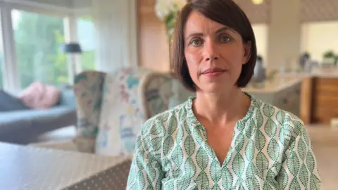 Angela O'Neill, a woman with brown, bobbed hair, looks into the camera.  She is wearing a white top patterned with green leaves.  There is a blurred background of a kitchen-diner behind her, with a large window on the left overlooking trees. 