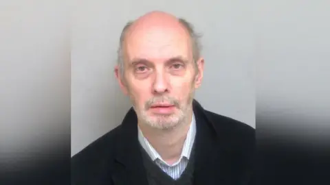 Essex Police David Pickthall wearing a black sweater and shirt. He is looking at the camera and has a grey beard.