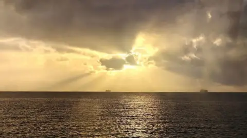 TomW A grey sea reflects yellow sunlight across it's choppy surface. The sun is hidden behind an equally grey cloud with beams of golden sunlight emanating across the sky