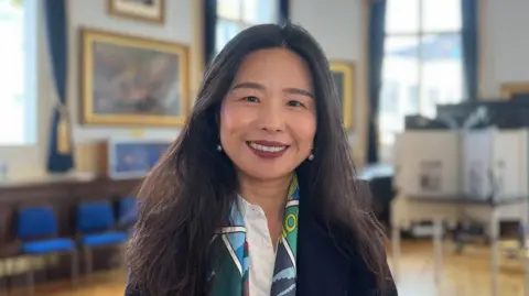 A woman with long, dark hair smiles in a large room. She's wearing a dark blazer over a white shirt, with a colourful and patterned green, blue, and white scarf. The room she's in includes large framed paintings on the wall and blue curtains around big windows.