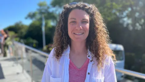 A women with curly brown hair smiling at the camera. She has sunglasses on top of her head. She is wearing a pink top with a white, linen style shirt over it with the buttons open.