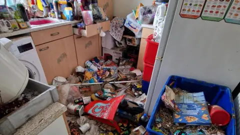 Ruth's kitchen is covered in rubbish. There are barely any visible surfaces, and there is food packaging waste everywhere. 