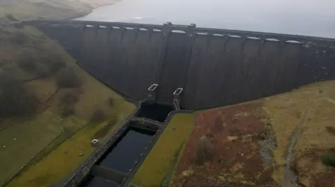 An aerial picture of the Clarven Dam imposed in BBC Mid Wales. The dam is made from gray stone and the arches at the top. It is surrounded by Banjar Misty Hills.