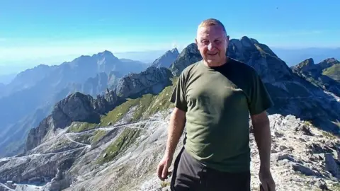 Rob Hann is pictured on a rocky ledge with many mountains in the background and bright blue skies