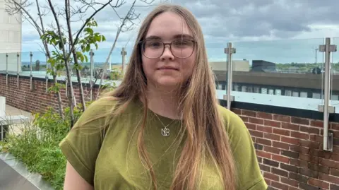A young woman stands in front of a low-brick wall and a planter. She has long brown hair and wears glasses, a green t-shirt and a silver necklace with a large pendant on it.