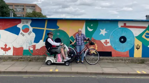 BBC Tony on his mobility scooter and Julie with her bicycle on Mill Road bridge